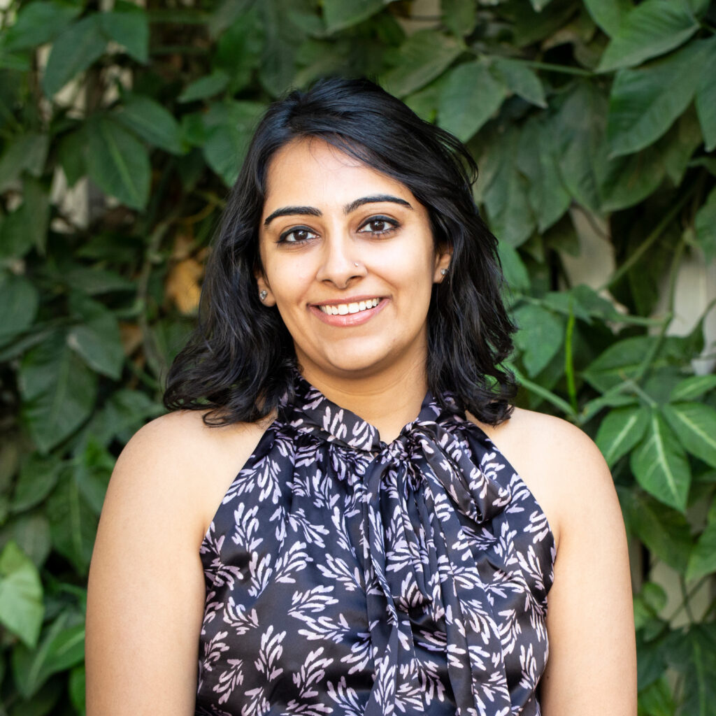 Portrait of a smiling woman with dark hair and a floral top, greenery background.