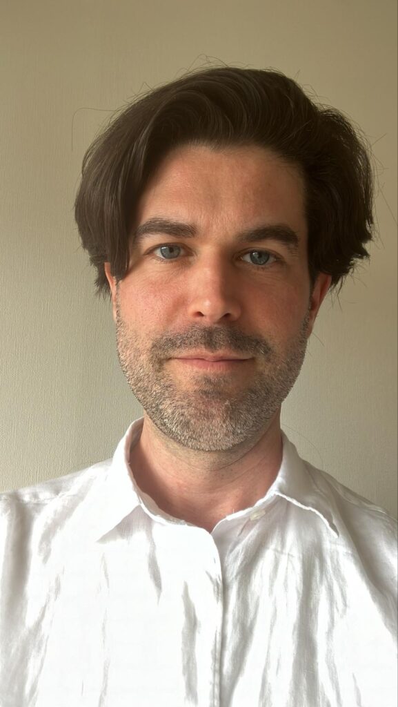 Close-up of a man with dark hair and blue eyes in a white shirt.