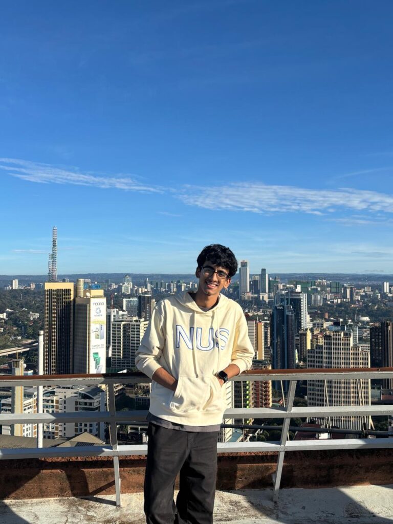 Young man standing on rooftop with city skyline in background under blue sky.
